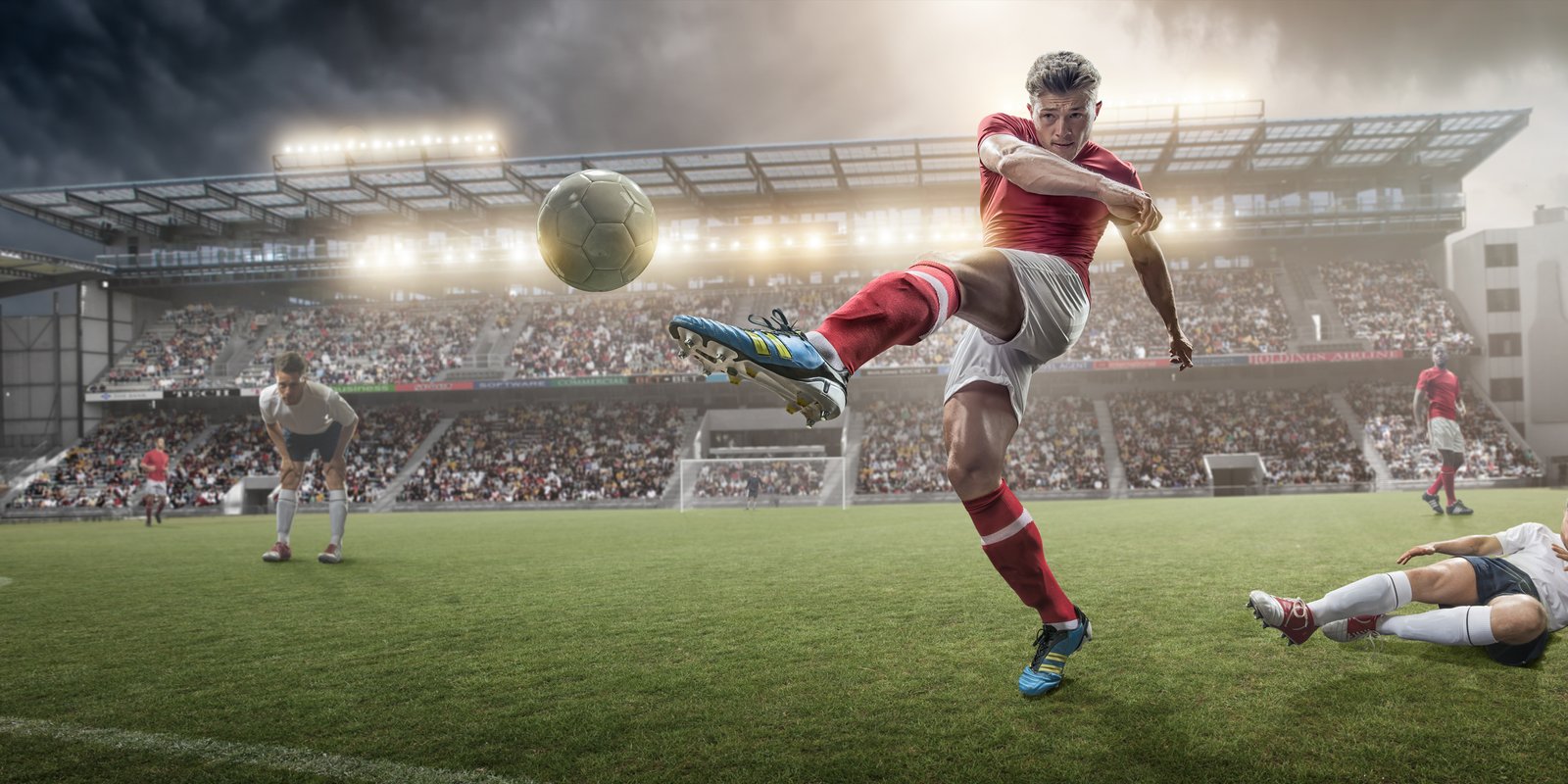 A mid action image of soccer player kicking football, following a sliding tackle challenge from a rival player. The action takes place on a soccer pitch on a generic outdoor floodlit football stadium full of spectators under a stormy evening sky at sunset. All players are wearing unbranded generic kit.